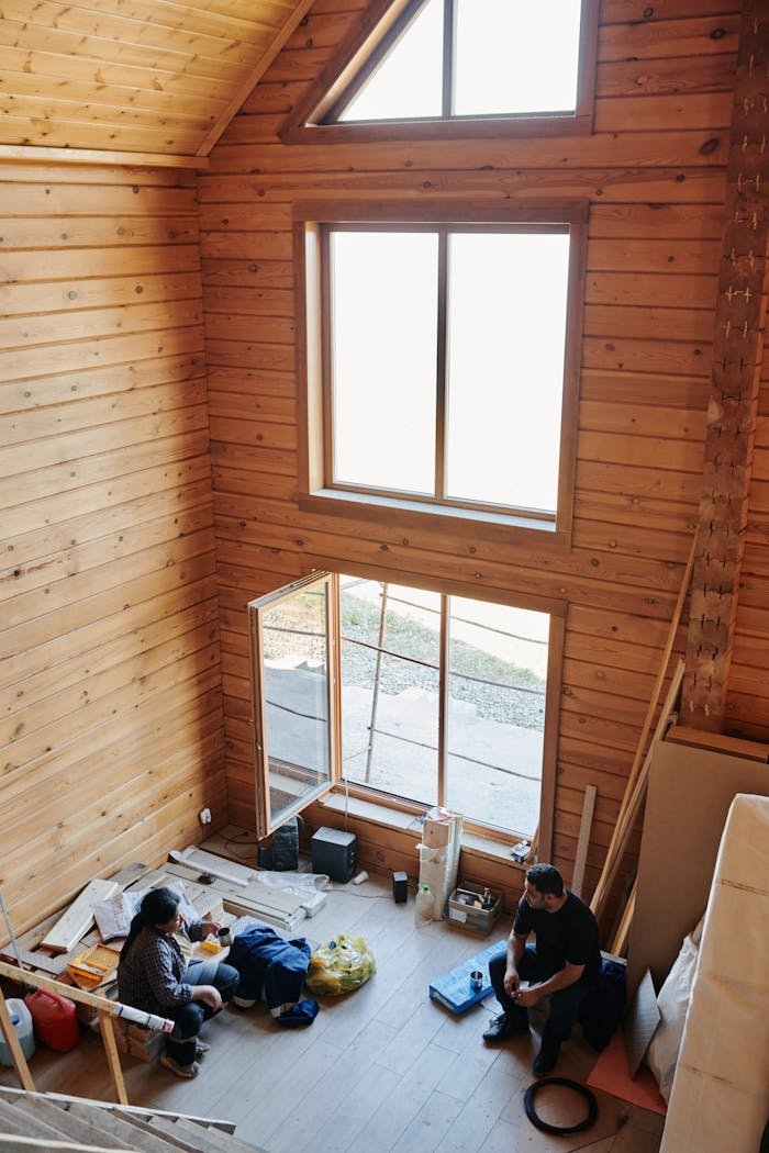 creative-01 Interior of a rustic wooden cabin with two people resting amid construction materials, capturing a moment of pause.